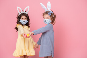 two girls in bunny ears headband and protective mask with colored eggs on pink background. Covid easter concept