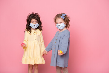 two girls in protective mask with colored eggs holding hands on pink background. Covid easter concept