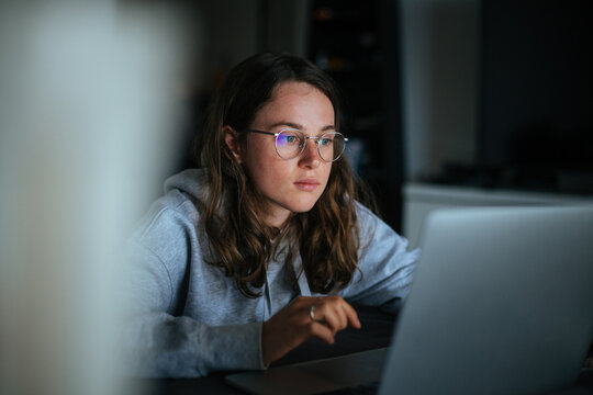 Millennial young woman sit at home and work on laptop.Home office concept, screen lit face from laptop, look at website and learn new skills from internet.Technology and information overload, big data