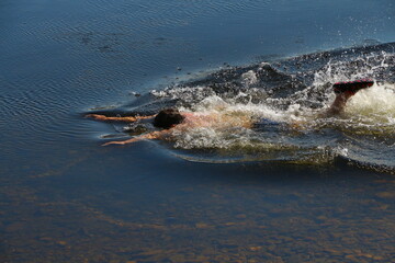 A man actively swims with his arms outstretched forward in transparent water with his head down, working with his foot in rubber shoes.Guy moves in calm water with splash and wave outdoors