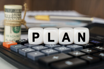There are cubes on the calculator that say - PLAN. Nearby out of focus - dollars, notebook and pen