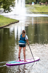 Woman paddling with SUP stand up paddle board in city canal near fountain. Active summer vacations with paddle board.