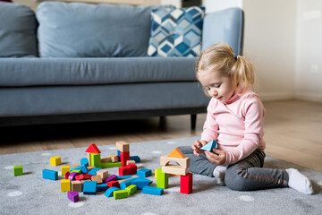 Cute Caucasian toddler girl playing with colorful wooden blocks on the floor of the living room at home. Activities with children indoor, early childhood development. Game with kids. Selective focus