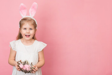 Obraz na płótnie Canvas Happy child girl wearing bunny rabbit ears with nest made of sticks full of painted eggs on pink background. Easter day children and fun concept.