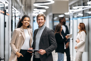 Portrait of two multiracial satisfied successful colleagues dressed in formal wear standing in a modern office, looking and friendly smiling at the camera