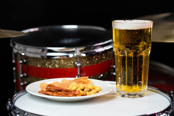 Glass of light beer with salty snacks on professional drum set closeup. Drumsticks, drums and cymbals, at live music rock concert, in the club stage, bar, or in recording studio