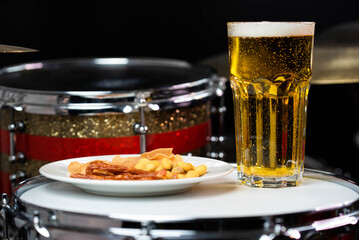 Glass of light beer with salty snacks on professional drum set closeup. Drumsticks, drums and cymbals, at live music rock concert, in the club stage, bar, or in recording studio
