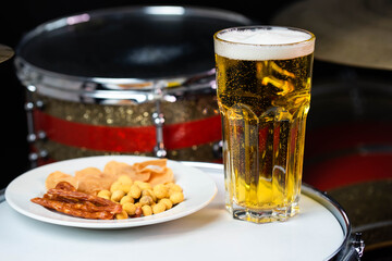 Glass of light beer with salty snacks on professional drum set closeup. Drumsticks, drums and cymbals, at live music rock concert, in the club stage, bar, or in recording studio