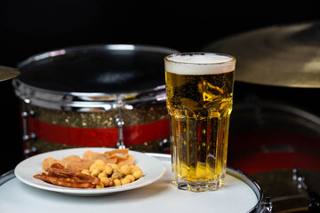 Glass of light beer with salty snacks on professional drum set closeup. Drumsticks, drums and cymbals, at live music rock concert, in the club stage, bar, or in recording studio