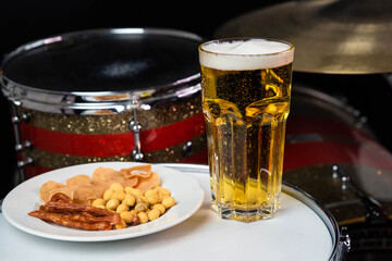 Glass of light beer with salty snacks on professional drum set closeup. Drumsticks, drums and cymbals, at live music rock concert, in the club stage, bar, or in recording studio