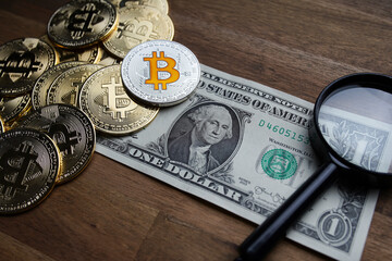 Pile of american dollars cash on brown wooden table. Next to it are several gold bitcoin digital cryptocurrency coins, a pen and a magnifying glass. Bank image and photo background.