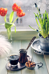 Oriental coffee in traditional copper coffee pot with flowers on window sill. Wooden windowsill with orange tulips and hyacinth flower pot. Cold rainy day in spring. Cozy scene, hygge concept.