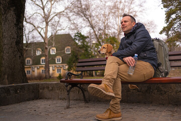 backpacker sitting on the bench at public park with his small yellow mix breed dog