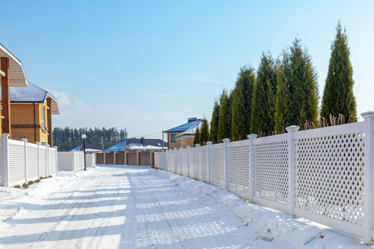 Snowy Road In A Cottage Village. White Plastic Fence In A Modern Cottage Village On A Clear Winter Day. Snow Drifts In Front Of A Vinyl Fence Against A Blue Sky