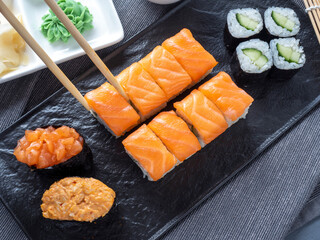 a variety of Japanese rolls and sushi on a textured black plate. Side view. Bamboo chopsticks holding a single roll
