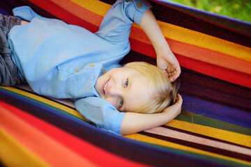 Cute little blond caucasian boy having fun with multicolored hammock in backyard or outdoor playground. Summer active leisure for kids. Child swinging and relaxing in hammock.