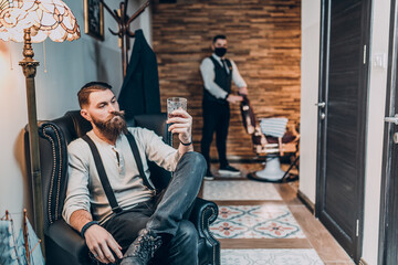Young good looking hipster man visiting barber shop and drinking whisky. Trendy and stylish beard...