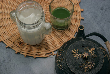 Horizontal image of crystal glass full of matcha tea mixed with sugar and honey while there is a cup tea next to a crystal glass full of ice milk on gray stone background. Matcha tea preparation 2021.