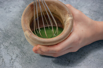 Horizontal image of clay bowl full of matcha tea with sugar and honey while beating with the kitchen rod on gray stone background. Matcha tea preparation 2021.