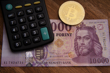 Hungarian forint 10,000 forint banknote Saint Istv&aacute;n King. Calculator on brown wooden table. Next to it is a gold bitcoin digital cryptocurrency coin and paper money. Bank image and photo.