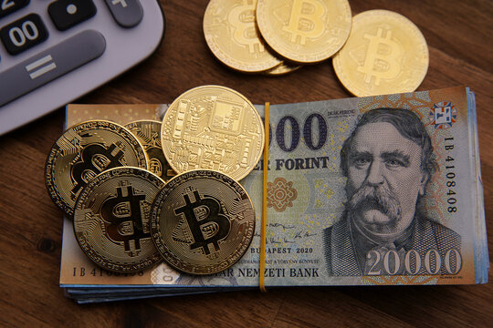 Hungarian forint 20,000 forint banknote stack. Brown wooden table. Next to it are several gold bitcoin digital cryptocurrency coins and a calculator. Bank image and photo.
