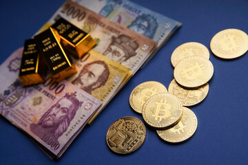 Various Hungarian banknotes, gold bars and bitcoin (BTC) coins on a blue table.