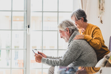 couple of two cheerful and happy mature and old people seniors using tablet and having fun sitting on the sofa at home together. Beauty retired pensioners using internet and surfing the net