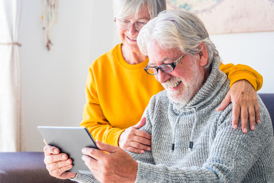 couple of two cheerful and happy mature and old people seniors using tablet and having fun sitting on the sofa at home together. Beauty retired pensioners using internet and surfing the net