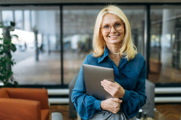 Joyful blonde-hair business woman in eyeglasses is looking at the camera, holding laptop in arms...