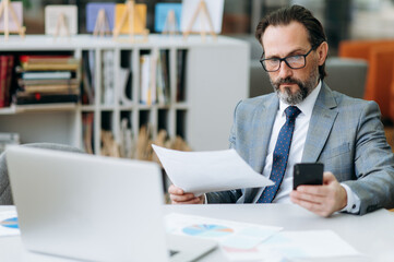 Fototapeta na wymiar Concentrated senior bearded businessman in eyeglasses uses smartphone, chatting with collegues. Serious businessman sitting at the desk, working on importnat project