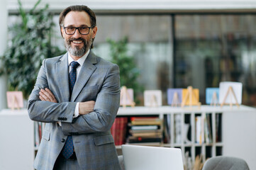 Portrait of a confident middle aged entrepreneur in stylish suit and eyeglasses standing in modern...