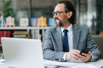 Joyful successful businessman sitting at the work desk, looks away and smiling. Satisfied mature male employee uses smartphone and laptop, thinking about new project