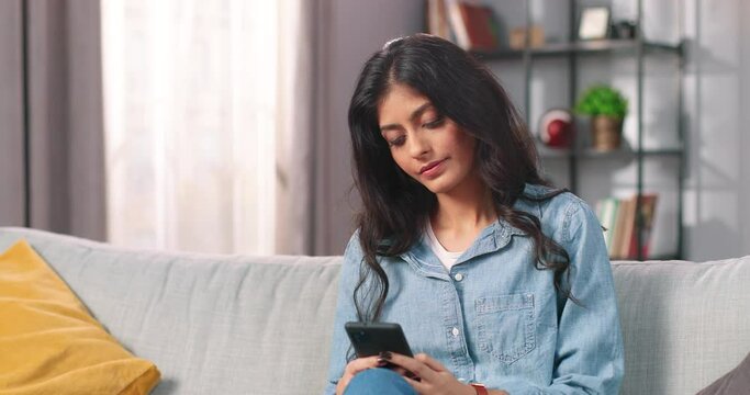 Indian joyful beautiful young woman typing on smartphone sitting on sofa in cozy room and smiling, pretty female searching internet on mobile phone using social network app, leisure concept, portrait