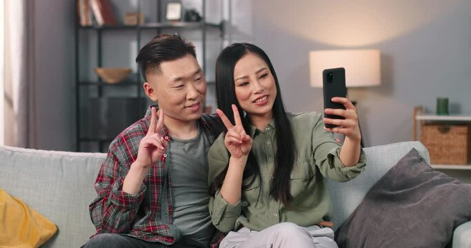 Close up portrait of cheerful Asian young couple man and woman taking selfie photos on smartphone at home in cozy room, posing to cellphone camera making gestures and smiling, leisure concept