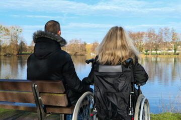 Concept of disabled person. A woman in a wheelchair outside in the nature in front of a lake. A man...