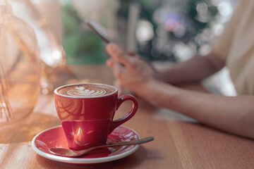Close up hot cappuccino, Latte coffee with blur hands of woman holding mobile, smart phone sitting in cafe. Morning time at coffee shop close up red coffee cup with blur hand hold phone vintage style