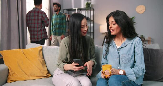 Portrait of mixed-race female friends sitting together at home on sofa and chatting typing on smartphone gadget while Asian and Hindu young guys speaking on background. Leisure concept