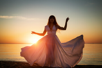Fototapeta na wymiar Beautiful female silhouette on the seashore in a white long dress at sunset