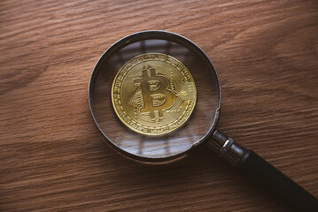 A magnifying glass magnify the replica of gold bitcoin on wooden background.