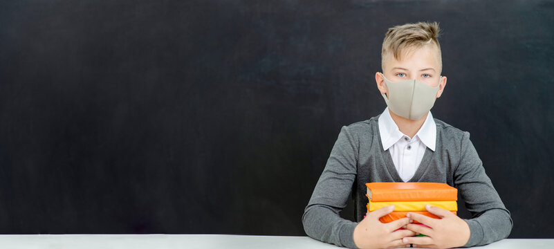 Teen schoolboy wearing mask sits with books at school near blackboard during corona virus and flu outbreak. Empty space for text
