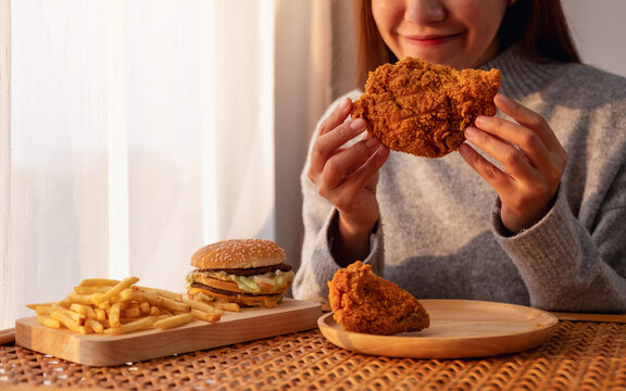 Closeup Image Of A Young Asian Woman Holding And Eating Fried Chicken With Hamburger And French Fries On The Table At Home