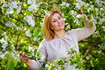 Portrait of a fat plump young woman with red hair and a blooming apple tree on a white floral background in a park on a spring day. Happy plus-size girl.