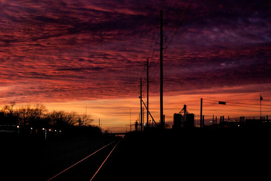 Textured Orange and Pink clouds at sunset behind a silhouetted grain elevator and power poles in a small midwest town with highlighted railroad tracks in the foreground leading off into the distance.