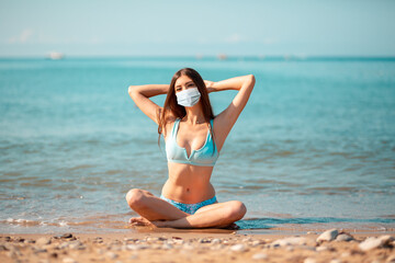 Fototapeta na wymiar Summer. A young caucasian woman in a protective mask, sitting crossed legs on the oceans beach. The concept of vacation during the coronavirus pandemic