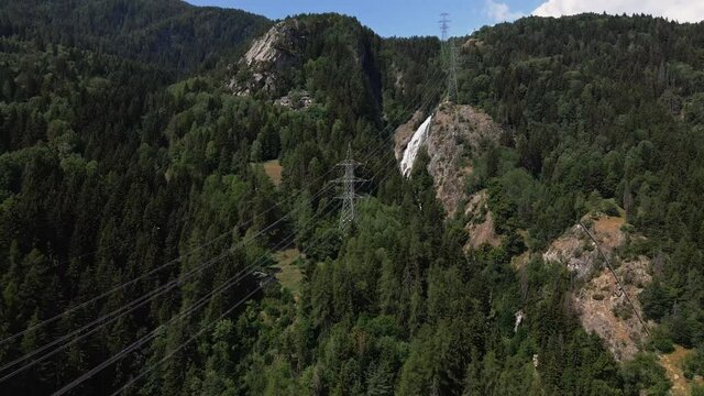 Power Cables Over The Italian Alps Surrounded By Green Woodland With Waterfall On The Background. aerial