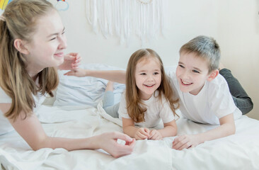 Obraz na płótnie Canvas The concept is happy brother and sister. Three children of different ages: a teenage girl, a little girl and a boy. Children in white T-shirts lie on the white bed and smile.