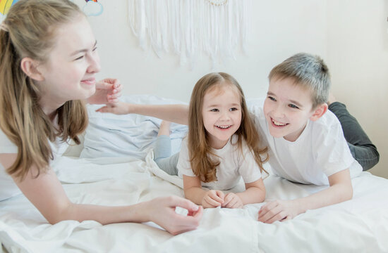 The concept is happy brother and sister. Three children of different ages: a teenage girl, a little girl and a boy. Children in white T-shirts lie on the white bed and smile.