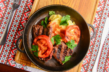 Close up of juicy beef steak served in a pan with bbq grilled tomatoes over traditional Ukrainian tablecloth.