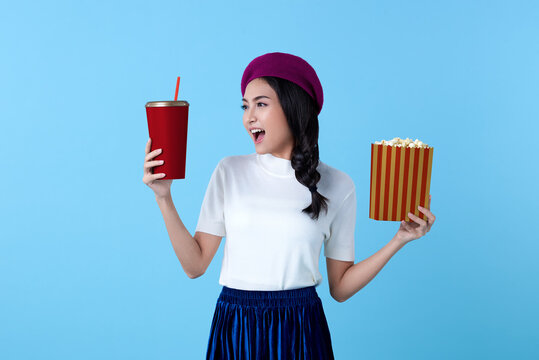 Excited Asian woman watching movie film holding popcorn and cup of soda isolated on bright blue background.