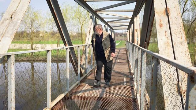 Disabled bearded senior man with walking stick moving slow and watching around on metal bridge over small river